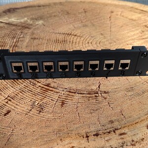 May include: A black network patch panel with eight RJ45 ports, numbered 1 to 8. The panel is designed for mounting in a standard 48.26 cm rack. The panel is made of metal and has mounting holes on each end.
