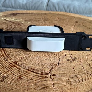 Könnte beinhalten: Eine schwarze Metall-Rack-Montagehalterung mit einem horizontalen rechteckigen Rahmen und kreisförmigen Befestigungslöchern an beiden Enden. Ein weißes, quadratisches elektronisches Gerät mit abgerundeten Ecken ist mittig fixiert.