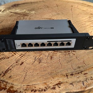 Może przedstawiać: Urządzenie sieciowe UniFi Switch 8 z szarą metalową obudową i czarnym wspornikiem do montażu w szafie rack. Przedni panel zawiera osiem portów Ethernet ponumerowanych od jednego do ośmiu, z logo UniFi i napisem Switch 8 na górnej powierzchni.