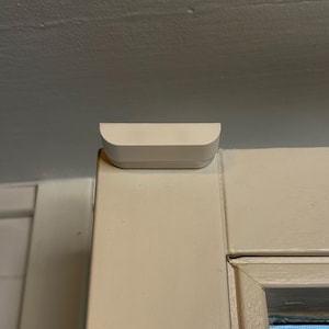 Op de afbeelding: Een klein, wit, rechthoekig apparaat met afgeronde randen is bovenop een wit deurkozijn gemonteerd. Het apparaat lijkt een sensor of een smart home-component te zijn, ontworpen om op te gaan in het deurkozijn.