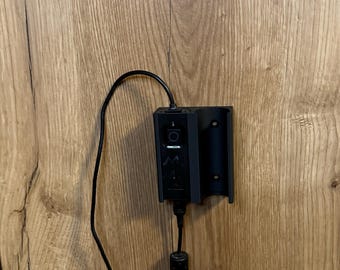 Può includere: Una stazione di ricarica nera con supporto a parete, cavo di alimentazione e un piccolo pannello di controllo. La stazione è montata su una superficie in legno con un motivo a venature naturali. Il cavo di alimentazione è collegato alla stazione.