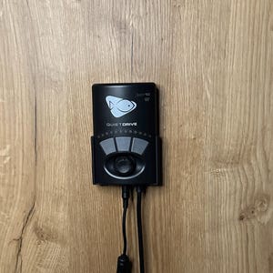 May include: Black Quiet Drive device with a fish logo and control buttons, mounted on a wooden surface. The text "Quiet Drive" is visible below the fish logo. Two black cords extend from the bottom of the device.