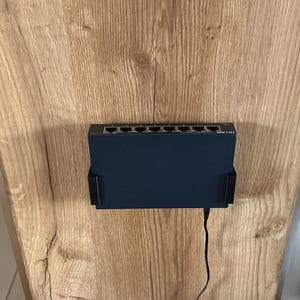Può includere: Uno switch di rete TP-Link nero montato in una staffa di plastica nera. Il dispositivo presenta otto porte Ethernet sul bordo superiore e un cavo di alimentazione che si estende dal basso. L'unità è fissata su una superficie in legno chiaro.