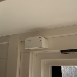 Può includere: Un dispositivo di sicurezza Ring bianco montato su un telaio di porta bianco. Il dispositivo ha il marchio "ring" e un design circolare. Il dispositivo è rettangolare con bordi arrotondati.