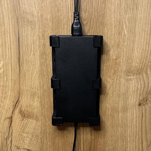 Könnte beinhalten: Eine rechteckige, schwarze Halterung für elektronische Geräte mit vier Eckclips, die vertikal montiert ist. Ein schwarzes Stromkabel geht von der oberen Mitte ab, und ein zweites schwarzes Kabel geht von der unteren Mitte des Geräts ab.
