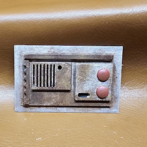 May include: A rectangular, weathered-looking panel with a textured surface. It features a series of vertical lines, two circular buttons, and a small rectangular opening. The panel is painted in shades of brown and gray, with a worn appearance.