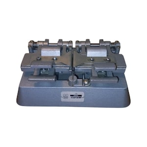 Puede incluir: Una herramienta industrial de metal gris con dos mecanismos de sujeción. La herramienta tiene una base rectangular y una serie de palancas y tornillos. Una pequeña etiqueta con texto es visible en la base. La herramienta parece ser utilizada para trabajos de precisión.