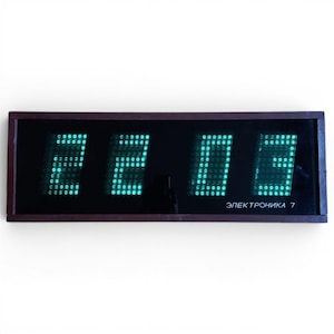 Pode incluir: Um relógio digital retangular com uma moldura castanha escura exibe a hora 22:03 em luzes LED verdes brilhantes. O mostrador do relógio é preto e o texto "ЭЛЕКТРОНИКА 7" é visível na parte inferior.