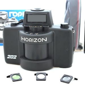 Könnte beinhalten: Schwarze Horizon 202 Panorama-Kamera mit dem Wort "HORIZON" in Weiß. Enthält ein Objektiv, eine Filmkartusche und drei Filter. Die Kamera befindet sich auf einer weißen Oberfläche mit einer blauen Box im Hintergrund.