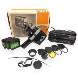 May include: A vintage black and silver movie camera with a lens, accompanied by its original cardboard box labeled KVARC-2, three film cassettes, a black leather carrying case, several circular lens filters, a lens hood, and a cable release.