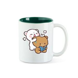 Könnte beinhalten: Eine weiße Keramiktasse mit grünem Innenraum. Die Tasse zeigt eine Cartoon-Illustration eines weißen Bären mit rosa Wangen und eines braunen Bären, der ein blaues Buch liest. Der weiße Bär beißt spielerisch in den Kopf des braunen Bären.