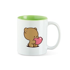 Könnte beinhalten: Weiße Keramiktasse mit grünem Rand. Die Tasse zeigt einen braunen Comic-Bären, der ein rosa Herz hält.