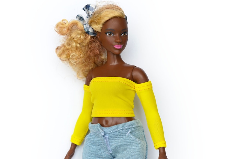 May include: A doll with brown skin and blonde curly hair wearing a yellow off-the-shoulder top with long sleeves and light blue jeans.