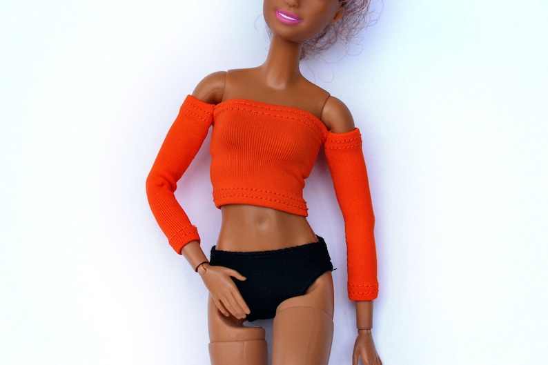 May include: A doll wearing an orange off-the-shoulder top with long sleeves and black bottoms.