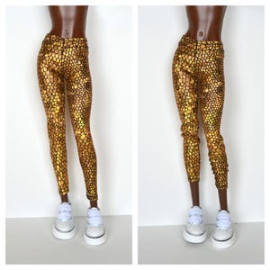 May include: Gold leggings with a scaly pattern and a black background. The leggings are worn by a doll with white sneakers.