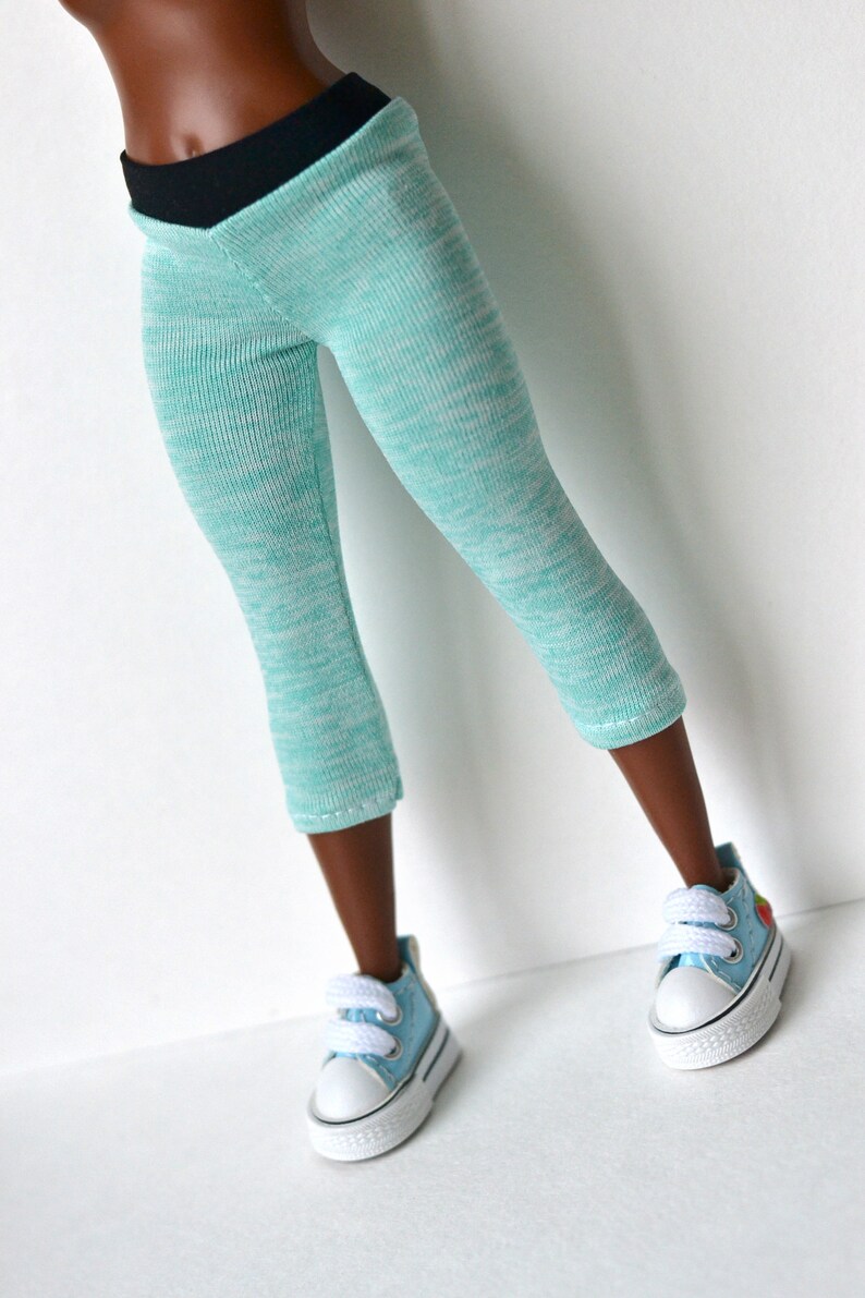 K&ouml;nnte beinhalten: Eine tealblaue Leggings mit schwarzem Bund. Die Leggings besteht aus einem weichen, dehnbaren Material und hat einen dezenten Melange-Effekt. Die Leggings wird von einer Puppe mit brauner Haut getragen und wird mit hellblauen Turnschuhen kombiniert.