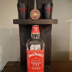 May include: A display featuring a Jack Daniel's Tennessee Fire bottle, a wooden shelf, two black shot glasses with a red emblem, and a firefighter coin. The bottle has a red label with white text. The shelf and supports are made of dark wood.