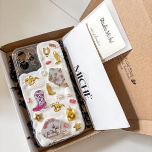 Könnte beinhalten: Eine transparente Handyhülle mit weißen Akzenten und goldenen Charms, darunter ein Cowboy-Stiefel, Sterne und ein Hufeisen. Die Hülle befindet sich in einer braunen Box mit dem Studio Miché-Logo auf einer Karte.