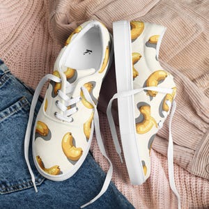 Puede incluir: Un par de zapatillas de lona blancas con un estampado repetido de ilustraciones de anacardos de dibujos animados en tonos amarillos y marrones. Los zapatos tienen cordones y suelas blancas. El fondo es una tela beige rosada y mezclilla.