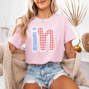 May include: A light pink t-shirt featuring the word "iM" in a playful design. The letter "i" is filled with a blue floral pattern, while the "M" is a red and white gingham check. The shirt is a casual, everyday wear item.