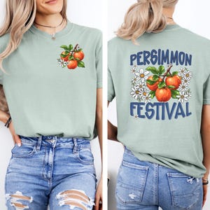 May include: A sage green t-shirt with a graphic of a persimmon branch with fruit and flowers on the front. The back of the shirt has the text "PERSIMMON FESTIVAL" in blue with a graphic of a persimmon branch with fruit and flowers.