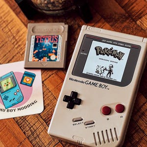 Puede incluir: Una consola Nintendo Game Boy vintage con el juego "Pokémon Version Rouge" en la pantalla. Un cartucho de juego "Tetris" y un posavasos con un diseño de Game Boy también están presentes. Un pequeño terrario de vidrio con una planta se encuentra en el fondo.