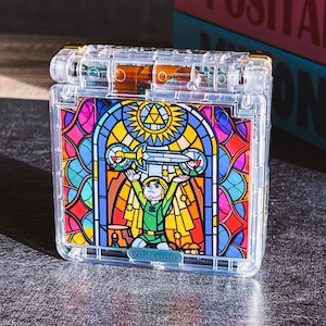 Op de afbeelding: Transparante plastic Nintendo Switch game cartridge case met een glas-in-lood ontwerp. De illustratie toont een personage in groene kleding met een zwaard, met een kleurrijke achtergrond van blauw, rood en geel. Het Nintendo-logo is zichtbaar.