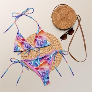 May include: A two-piece bikini set with a colorful abstract floral pattern in shades of blue, purple, orange, and pink. The bikini top has halter straps, and the bottoms have tie sides. A round woven bag and sunglasses are also in the image.