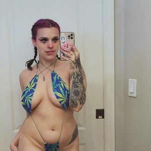 May include: A woman wearing a green and blue marijuana leaf print swimsuit with thin straps. The swimsuit is a one-piece with a low cut and a high leg. The woman has pink hair and is wearing makeup. She is standing in front of a mirror and holding a phone.
