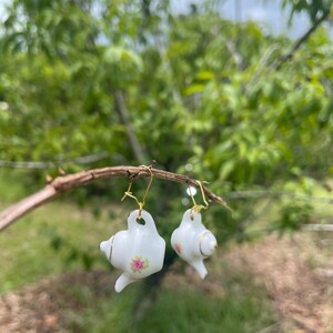 May include: White porcelain teapot earrings with gold-colored accents and pink floral details. The earrings are suspended from gold-colored ear wires. The background is a blurred view of green foliage.