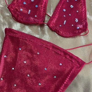 Peut inclure: Un ensemble de bikini rouge orn&eacute; de paillettes. Le haut triangle et le bas sont orn&eacute;s de multiples strass iris&eacute;s. Le tissu a un l&eacute;ger scintillement et les bords sont soigneusement cousus. Le bikini est pr&eacute;sent&eacute; sur une surface dor&eacute;e textur&eacute;e.