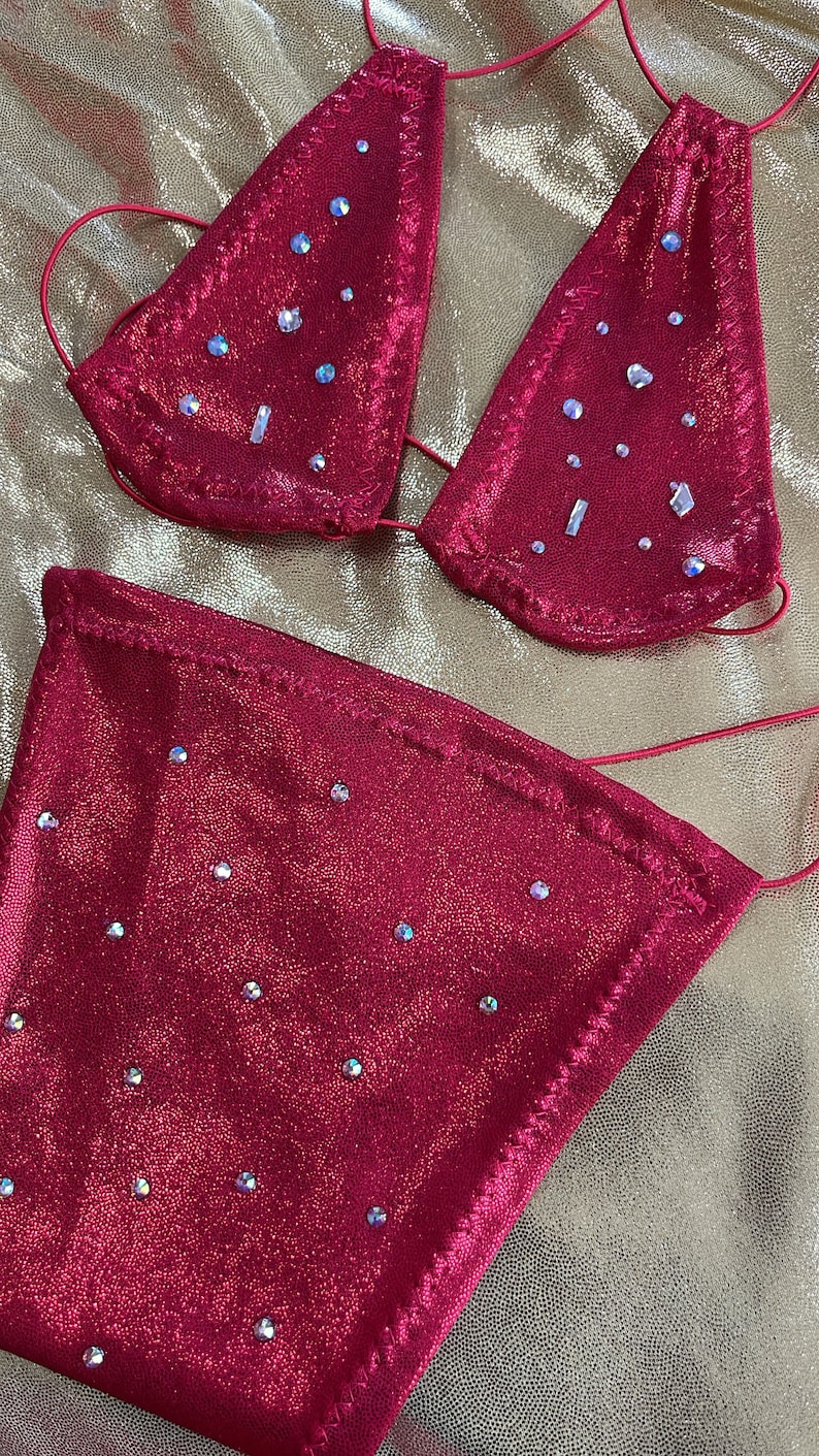 Peut inclure: Un ensemble de bikini rouge orn&eacute; de paillettes. Le haut triangle et le bas sont orn&eacute;s de multiples strass iris&eacute;s. Le tissu a un l&eacute;ger scintillement et les bords sont soigneusement cousus. Le bikini est pr&eacute;sent&eacute; sur une surface dor&eacute;e textur&eacute;e.