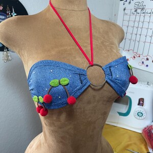 May include: A denim-style bikini top with a red halter strap and a silver ring detail. The top is embellished with red cherry accents and green leaves. Rhinestones are scattered across the fabric.