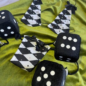 May include: An assortment of dice-themed accessories, including black dice with white dots and diamond-patterned triangle-shaped ornaments. The accessories are adorned with black bows and small, sparkling embellishments, set against a green velvet backdrop.