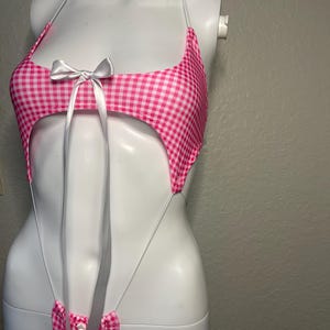 May include: Pink and white gingham one-piece swimsuit with a white bow at the bust and thin white straps.
