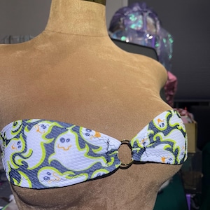 May include: A strapless bikini top featuring a repeating pattern of white cartoon ghosts with neon green outlines on a dark blue background. The top is connected with a gold-colored ring.