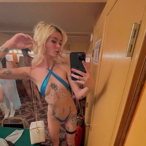 May include: A woman with blonde hair is wearing a blue halter top and a black garter belt. She has several tattoos on her arms, chest, and legs. She is holding a smartphone in her right hand and taking a selfie in a mirror. A small white box with a gold chain is hanging from her shoulder.