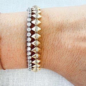 May include: Two bracelets are shown. One is gold with diamond-shaped accents and small diamonds. The other is silver with small, round diamonds. The bracelets are stacked on top of each other.