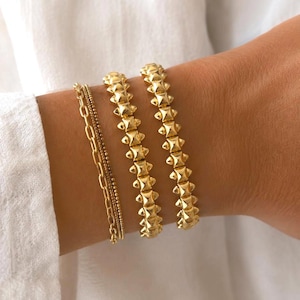 Könnte beinhalten: Ein Set aus vier goldfarbenen Armbändern, bestehend aus zwei feinen Kettenarmbändern und zwei passenden Armbändern mit einem sich wiederholenden Muster aus kleinen, spitzen pyramidenförmigen Nieten in einer Reihe.