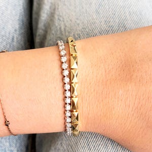 May include: A close-up of three bracelets. One is a delicate gold chain with a small charm. The second is a tennis bracelet with small, round, clear stones. The third is a gold bracelet with square, pyramid-shaped accents.