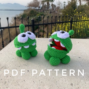 May include: Two green crocheted plush toys shaped like a monster with a large mouth. The toys have white eyes and teeth. The text "PDF PATTERN" is printed on the surface below the toys.