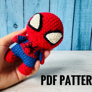 Könnte beinhalten: Ein rotes und blaues gehäkeltes Spiderman Amigurumi-Spielzeug mit schwarzen Augen und einem Spinnen-Symbol auf der Brust. Das Spielzeug wird in einer Hand gehalten. Der Text "PDF PATTERN" ist am unteren Rand des Bildes sichtbar.