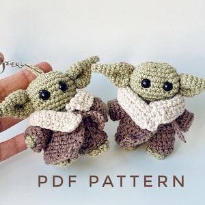 Könnte beinhalten: Zwei gehäkelte Yoda-Schlüsselanhänger aus Grün und Braun mit schwarzen Augen. Die Figuren tragen braune Mäntel mit weißen Kragen. Der Text "PDF PATTERN" befindet sich am unteren Rand des Bildes.