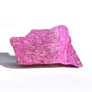May include: A vibrant, rough-hewn, pink mineral specimen. The rock has a textured surface with subtle variations in color and tone. The specimen is set against a white background, highlighting its natural beauty.