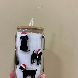 May include: A glass tumbler with a bamboo lid and a clear glass straw. The tumbler has a white background with black silhouettes of dogs wearing red Santa hats.