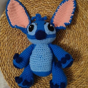May include: A handmade blue crochet plush toy with large ears, black eyes, and a dark blue nose. The inside of the ears is pink. The toy is sitting on a woven, circular surface. The plush toy is a character from a popular animated film.