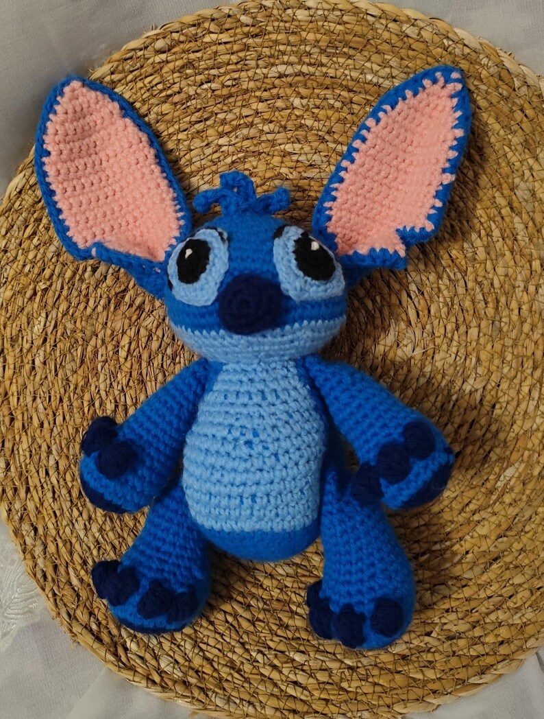 May include: A handmade blue crochet plush toy with large ears, black eyes, and a dark blue nose. The inside of the ears is pink. The toy is sitting on a woven, circular surface. The plush toy is a character from a popular animated film.
