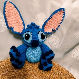 May include: A handmade blue crochet Stitch doll with large ears, black eyes, and dark blue accents. The plush toy is sitting on a woven surface, showcasing its detailed craftsmanship and vibrant colours.