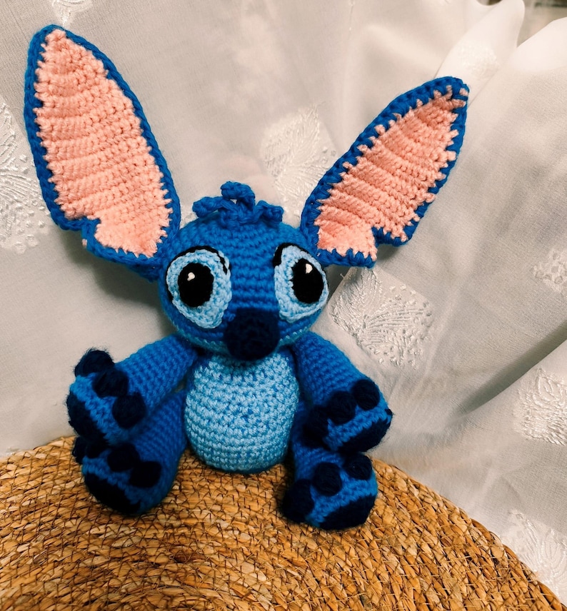 May include: A handmade blue crochet Stitch doll with large ears, black eyes, and dark blue accents. The plush toy is sitting on a woven surface, showcasing its detailed craftsmanship and vibrant colours.