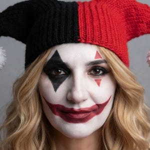 May include: A two-toned jester hat featuring a black side and a red side, each ending in a white pom-pom. The hat is worn over blonde hair, paired with white face paint, a black diamond eye marking, red triangle accents, and red lipstick.